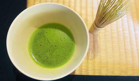 Bright green ceremonial matcha with frothy crema served in a traditional bowl alongside a bamboo chasen on a tatami mat.
