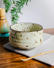 Japanese ceramic chawan with speckled glaze, bamboo whisk (chasen) and chashaku on a wooden table.