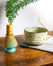 Speckled Japanese chawan with bamboo chashaku; bamboo whisk on a turquoise stand (kusenaoshi) on wooden table.