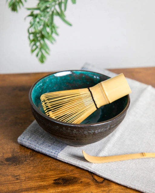 Japanese chawan with teal glaze holding a bamboo whisk (chasen); bamboo chashaku on cloth on a wooden table.