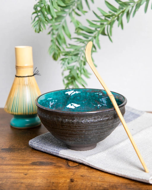 Japanese chawan with teal glaze, bamboo chashaku upright on rim; bamboo whisk on turquoise stand in the background.