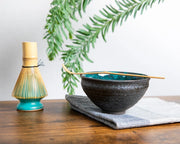Teal-glazed Japanese chawan on cloth with bamboo chashaku across rim; bamboo whisk on turquoise stand (kusenaoshi) nearby.