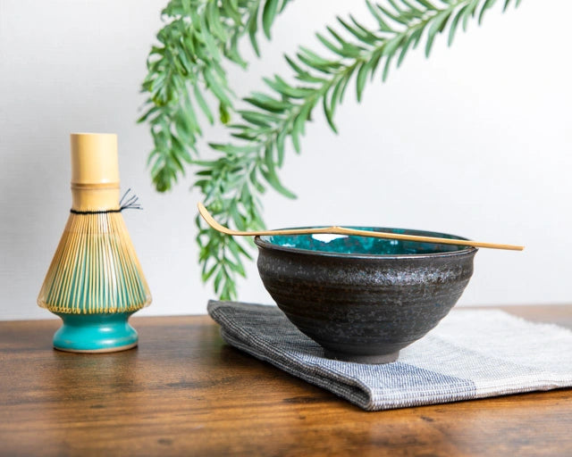 Teal-glazed Japanese chawan on cloth with bamboo chashaku across rim; bamboo whisk on turquoise stand (kusenaoshi) nearby.