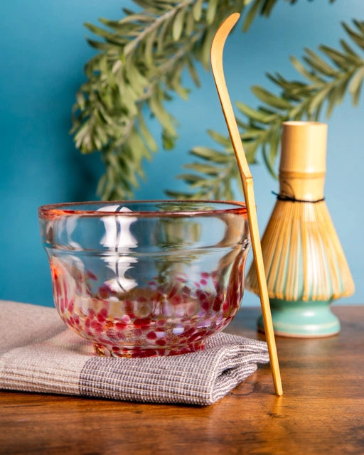 Japanese glass chawan with red accents on cloth, bamboo chashaku upright; bamboo whisk (chasen) on turquoise stand in background.