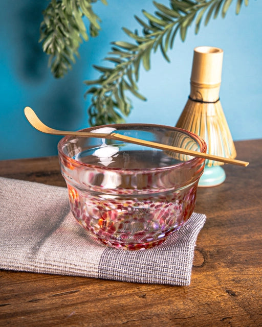 Japanese glass chawan with red accents on folded cloth, bamboo chashaku resting across the rim; whisk in background.