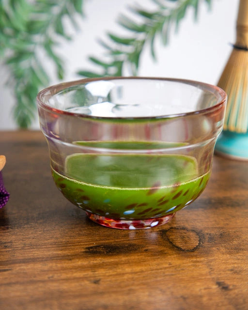 Glass chawan with red accents holding whisked green matcha on a wooden table; whisk in background.