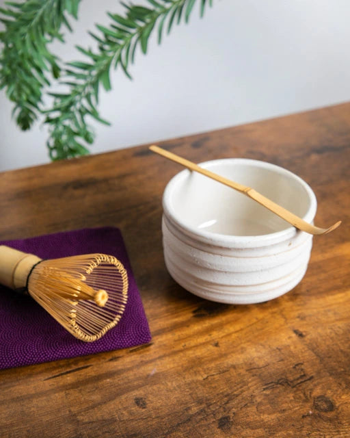 White ceramic chawan with bamboo chashaku resting on rim; bamboo whisk on purple cloth on a wooden table.