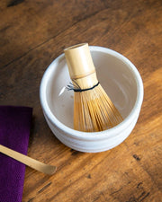 White ceramic chawan with bamboo whisk (chasen) inside; bamboo chashaku and purple cloth on a wooden table.