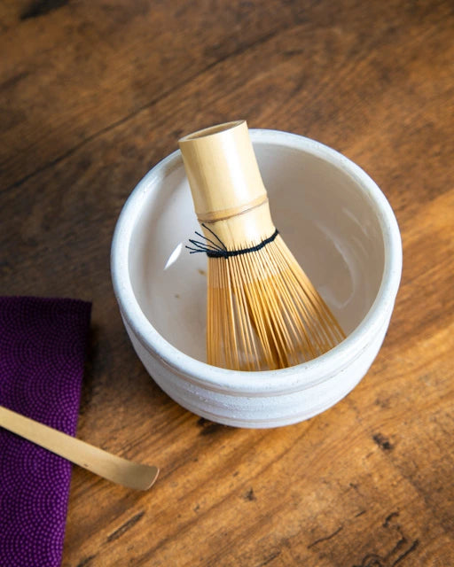 White ceramic chawan with bamboo whisk (chasen) inside; bamboo chashaku and purple cloth on a wooden table.