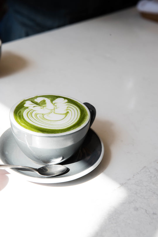 A cup of matcha latte with swan latte art in a grey ceramic mug on a white marble table with a spoon on the saucer.
