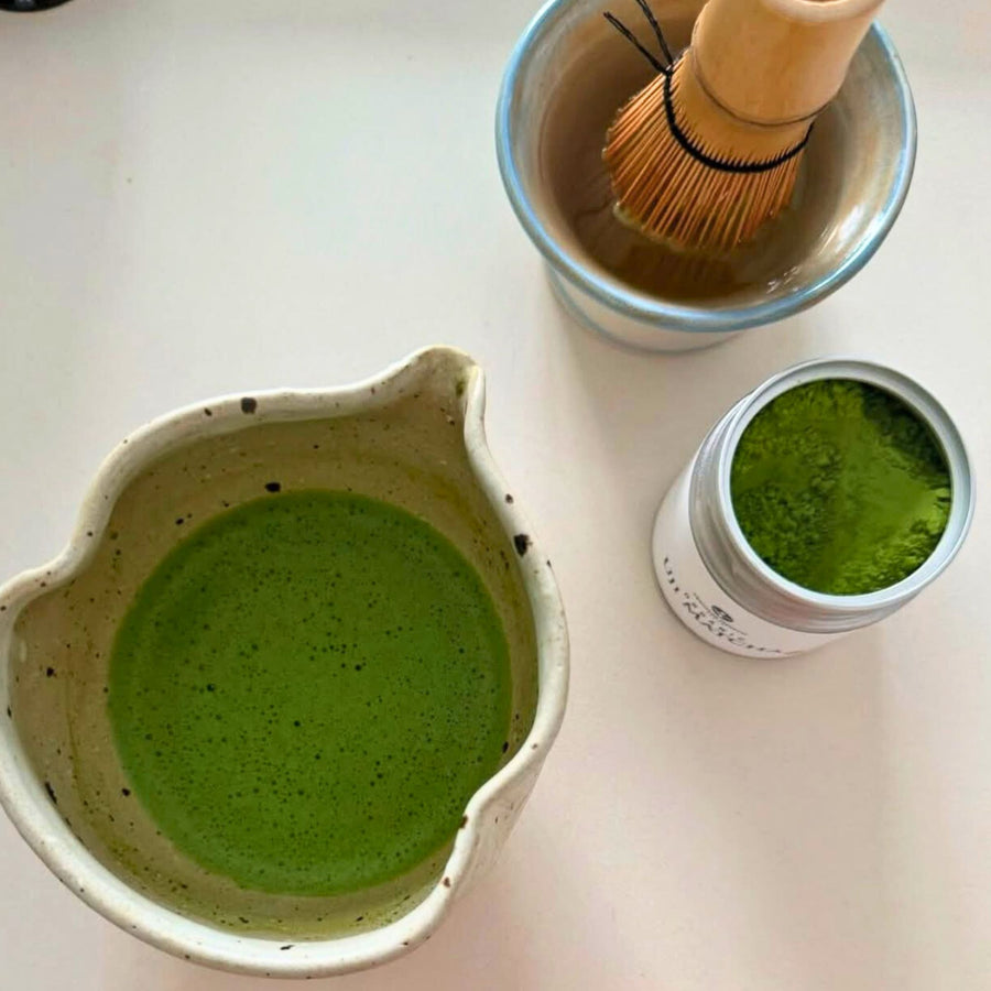 Overhead view of prepared organic matcha in a ceramic bowl, with a bamboo whisk soaking and open tin of vibrant green matcha powder.