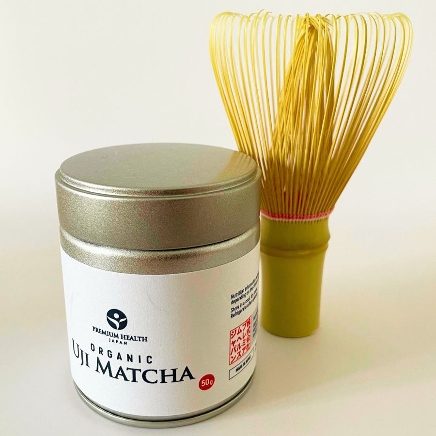 50g tin of Premium Health Japan Organic Uji Matcha next to a traditional bamboo whisk (chasen) on a light background.