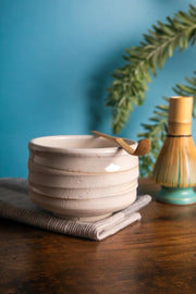 White ceramic chawan with bamboo chashaku on rim; bamboo whisk on turquoise stand (kusenaoshi) against a blue backdrop.