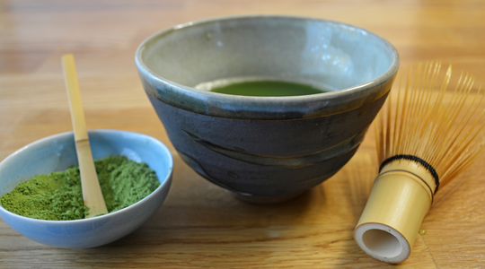 Matcha green tea powder, bowl of matcha tea, chashaku, and bamboo whisk on a table