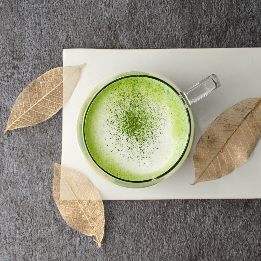 Top-down view of an Asahi matcha latte dusted with matcha powder, in a glass mug on a white board with gold skeleton leaves