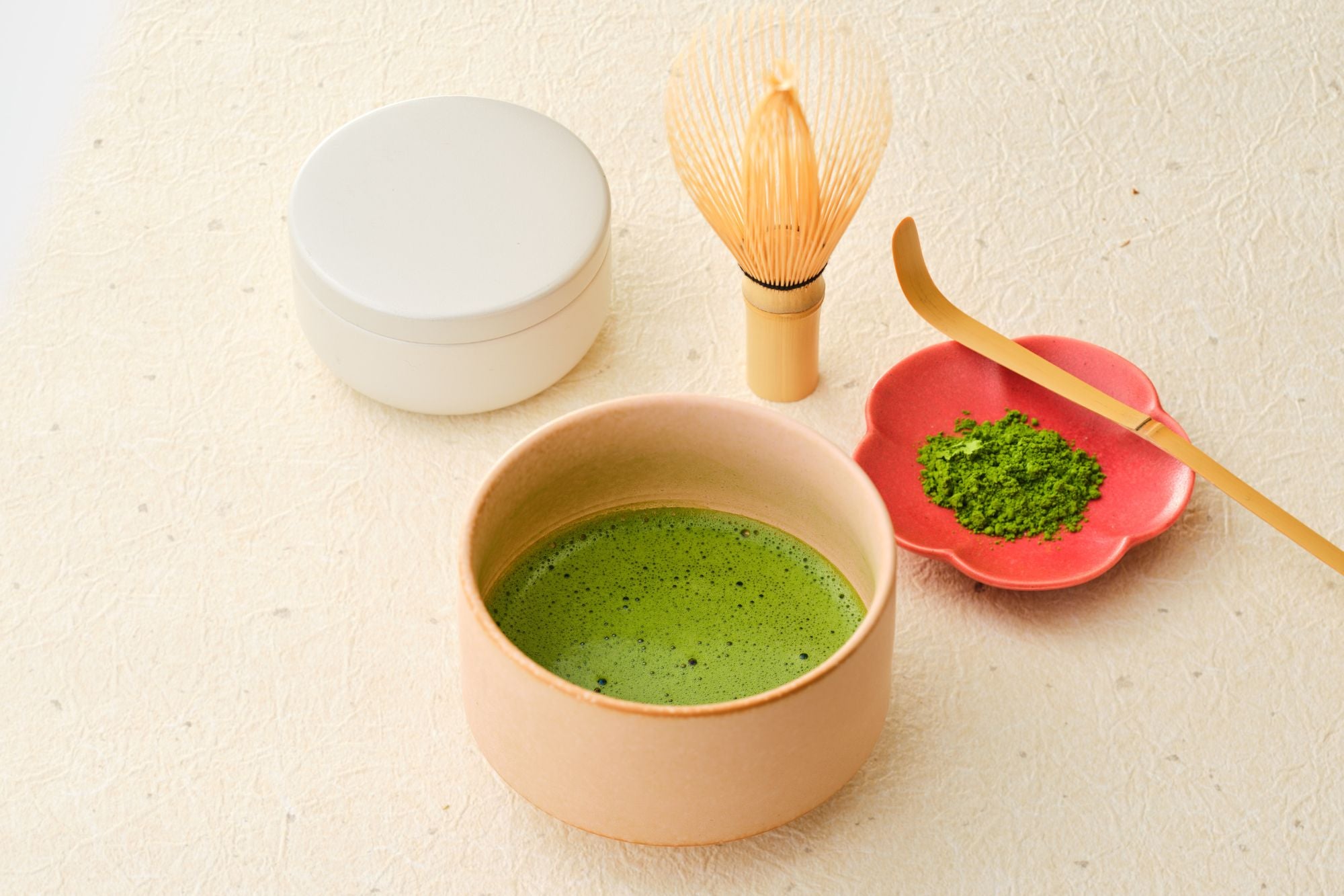 First spring harvest Uji matcha with a white tin, whisked bowl of matcha, bamboo chasen and chashaku, and a red flower dish of green powder on cream washi paper