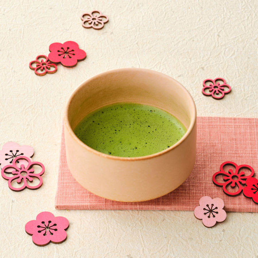 Whisked ceremonial grade Uji matcha from the first spring harvest in a peach ceramic chawan on a pink cloth, surrounded by wooden plum blossom ornaments