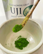 Close-up of vibrant ceremonial grade Uji matcha powder on a bamboo chashaku above a matcha bowl, with the Premium Health Japan 100 g pouch in the background.