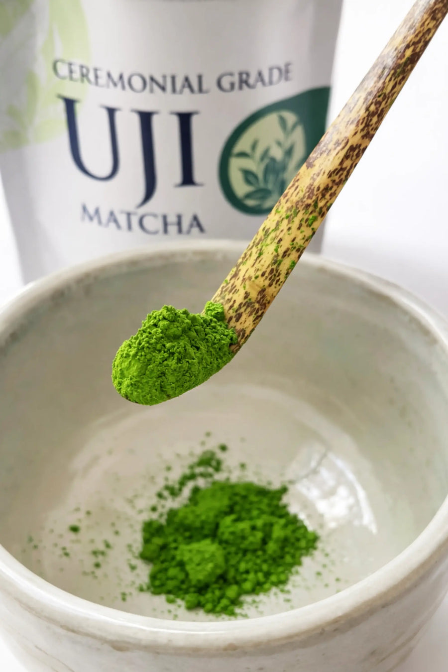 Close-up of vibrant ceremonial grade Uji matcha powder on a bamboo chashaku above a matcha bowl, with the Premium Health Japan 100 g pouch in the background.