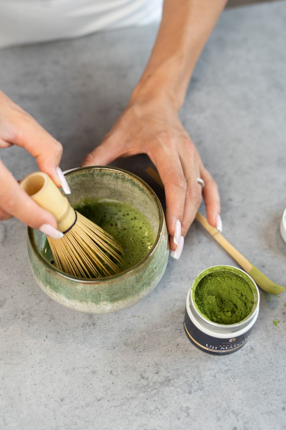 An open can of matcha green tea powder next to a bowl of matcha being made with a bamboo whisk