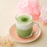Hot matcha latte made with first spring harvest ceremonial Uji matcha in a glass mug on a pink saucer, with cherry blossoms behind