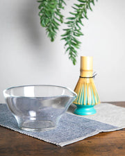 Clear glass chawan on layered cloth with bamboo whisk (chasen) in background, tabletop styling.