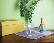 Clear glass chawan with green gift box and bamboo whisk (chasen) on cloth, styled on a wooden table.