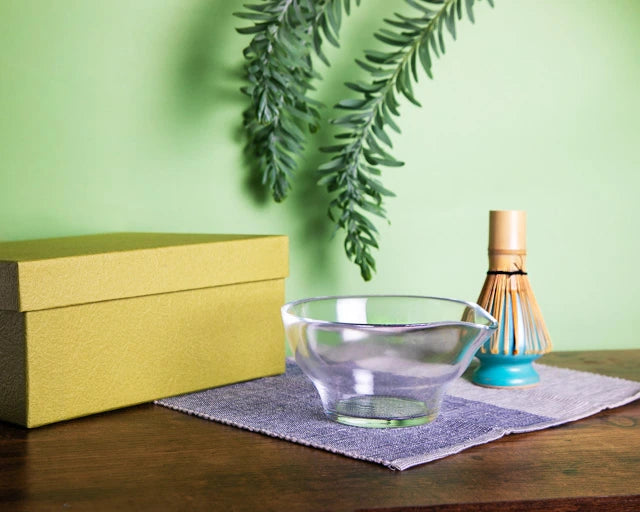 Clear glass chawan with green gift box and bamboo whisk (chasen) on cloth, styled on a wooden table.