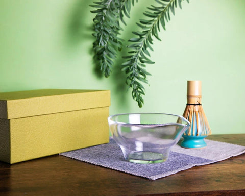 Clear glass chawan with green gift box and bamboo whisk (chasen) on cloth, styled on a wooden table.