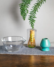 Clear glass chawan with bamboo whisk in tube packaging and turquoise whisk stand (kusenaoshi) on cloth, tabletop styling.