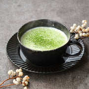Hot matcha latte made with Uji Matcha Gokō × Samidori blend, matcha dusted over the foam in a black ceramic cup and saucer with white berry branches