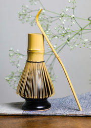 Handcrafted bamboo matcha whisk resting on a black whisk holder with a chashaku beside it, symbolizing traditional Japanese tea ceremony craftsmanship.