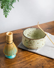 Speckled ceramic chawan with bamboo chashaku and bamboo whisk on stand (kusenaoshi) on a wooden table.
