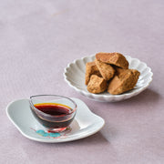 Japanese kuromitsu black sugar syrup in a small glass dish on a porcelain plate, with a scalloped dish of rough-cut Okinawa kokuto brown sugar behind