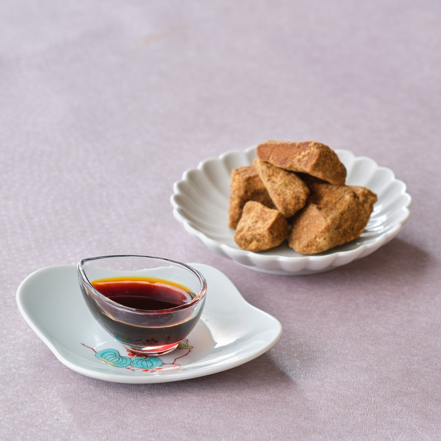Japanese kuromitsu black sugar syrup in a small glass dish on a porcelain plate, with a scalloped dish of rough-cut Okinawa kokuto brown sugar behind