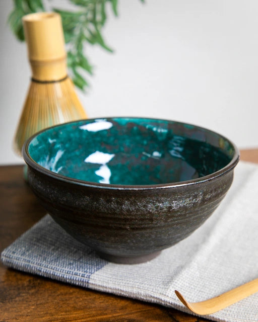 Japanese matcha bowl (chawan) with teal interior glaze, bamboo chashaku in front and chasen in background on cloth.