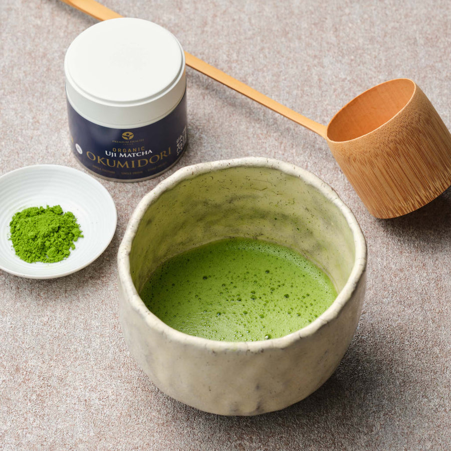 JAS Organic Uji Matcha Okumidori single-cultivar 30g tin beside a whisked bowl of matcha in a raku chawan, matcha powder on a white dish and a bamboo hishaku ladle