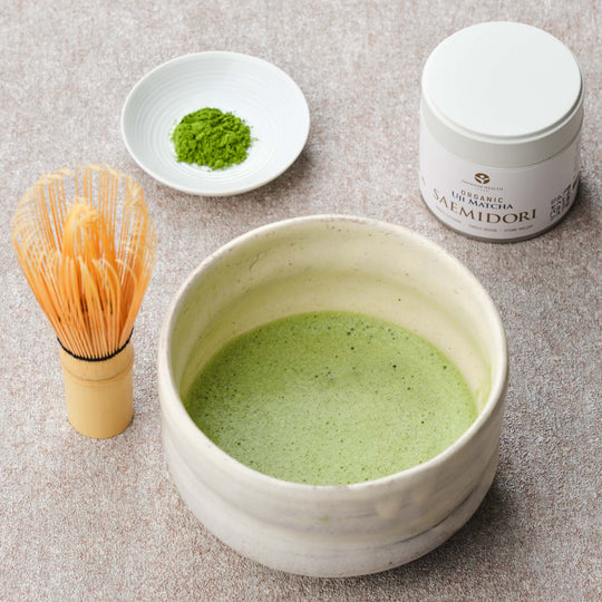 JAS Organic Uji Matcha Saemidori single-cultivar 30g tin with a whisked bowl of matcha in a cream chawan, bamboo chasen whisk and matcha powder on a white dish