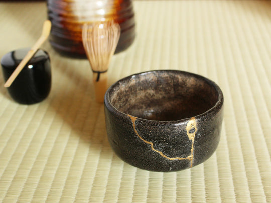Kintsugi tea bowl repaired with gold, placed on tatami mats alongside traditional tea ceremony utensils.
