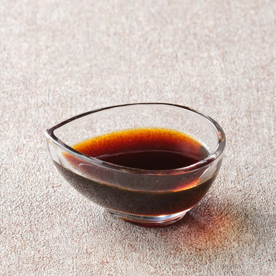 Close-up of authentic Japanese kokuto mitsu brown sugar syrup, glossy amber-black, in a clear glass dish on a stone surface