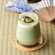 Okinawa brown sugar syrup being drizzled over an iced matcha latte in a glass on a bamboo coaster, with a Japanese folding fan behind