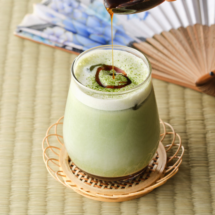 Okinawa brown sugar syrup being drizzled over an iced matcha latte in a glass on a bamboo coaster, with a Japanese folding fan behind