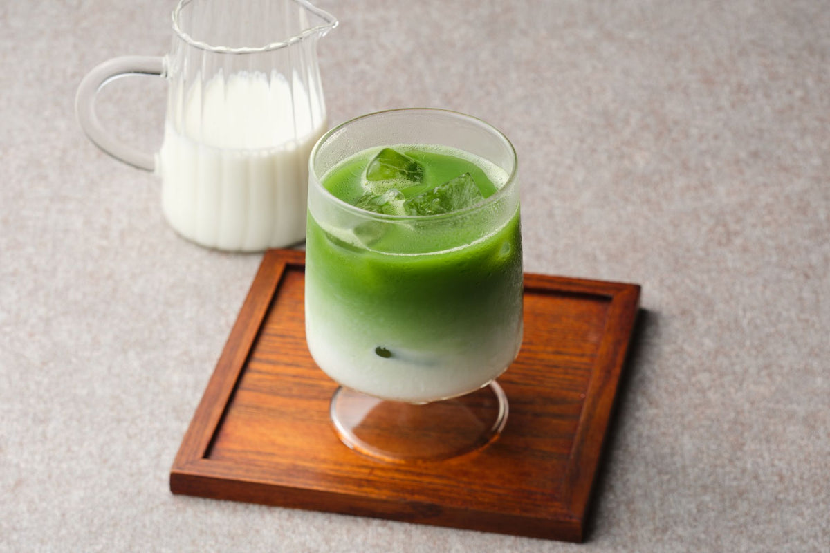 Iced matcha latte layered over milk in a glass on a wooden tray, with a jug of milk behind on a beige surface