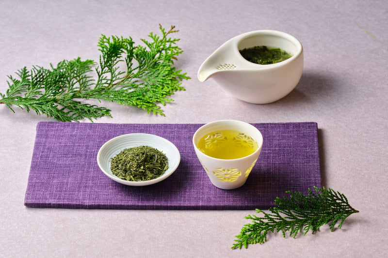 Loose-leaf Japanese sencha in a white dish beside a brewed cup and a kyusu teapot on a purple cloth with cedar sprigs