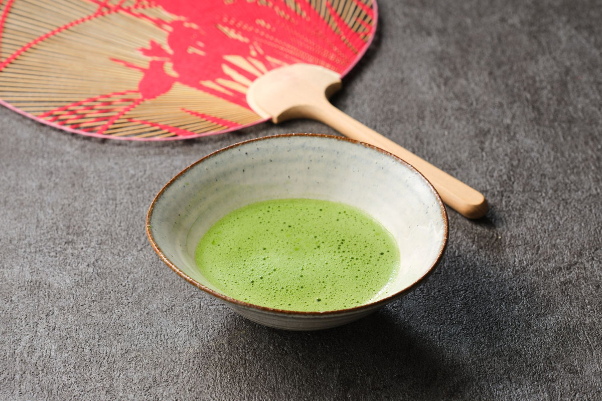 Freshly whisked usucha matcha in a rustic ceramic chawan with a bamboo chashaku and a red paper fan on dark stone