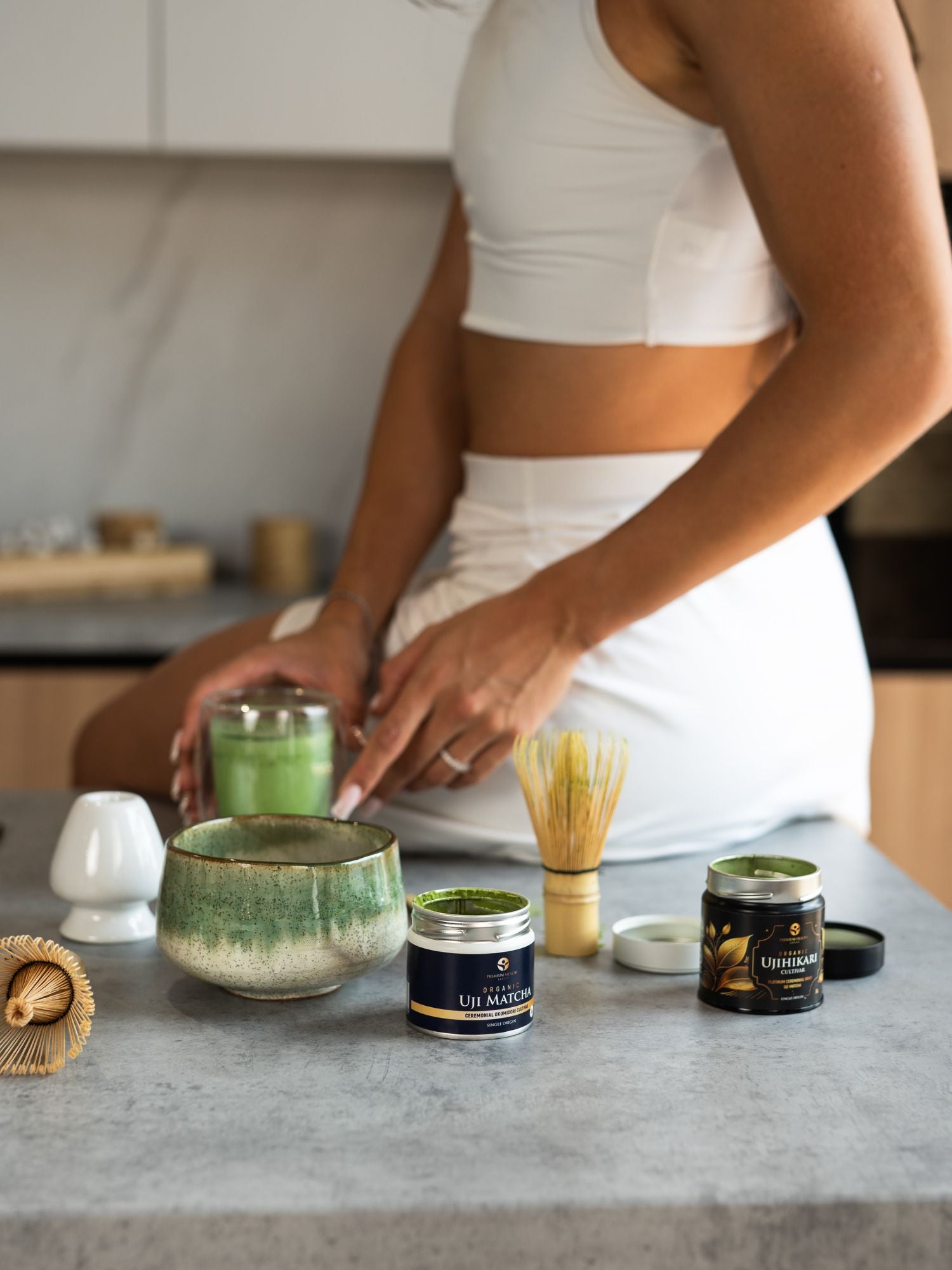 Woman sitting on bench drinking a matcha latte, with a matcha bowl, Uji matcha products, and a bamboo matcha whisk beside her