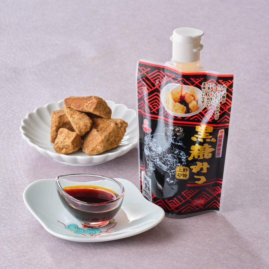 Okinawa brown sugar syrup (kokuto mitsu) 200g squeeze pouch beside a glass dish of dark syrup and a plate of raw Okinawan kokuto black sugar chunks