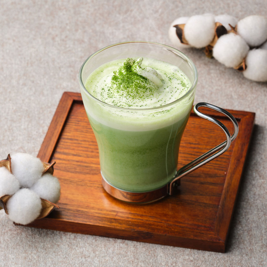 Hot matcha latte made with organic single-cultivar Okumidori Uji matcha, topped with whipped cream and dusted matcha, in a glass mug on a wooden tray with cotton bolls