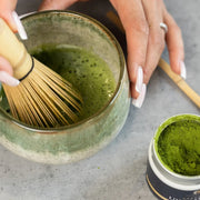 Whisking Okumidori organic Uji matcha into frothy usucha with a bamboo chasen, beside an open tin of vivid green Kyoto matcha powder