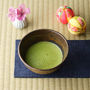 Whisked organic ceremonial grade matcha in a rustic dark raku chawan on an indigo cloth and tatami mat, with pink flowers and temari balls